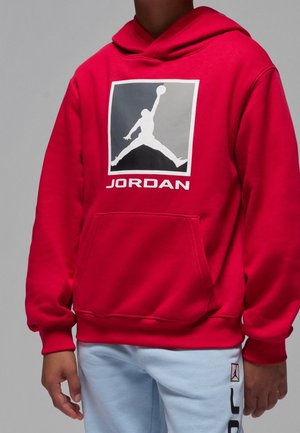 Person iført en rød hættetrøje med et sort, gråt og hvidt basketballlogo og teksten "JORDAN", kombineret med lyseblå bukser med logopatch.