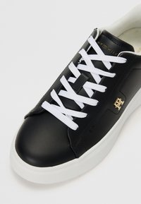 Black leather sneaker with white laces, padded tongue, and a gold accent logo. Features a rounded toe and chunky white rubber sole.