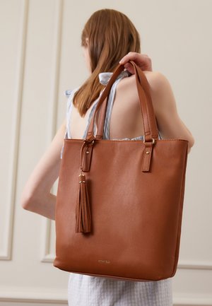 Brun læder totebag med dobbelte stropper, en dekorativ frynse og subtile guldfinish. Enkel rektangulær form med glat tekstur.
