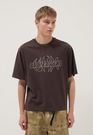 Joven con el pelo corto y castaño lleva una camiseta holgada de color marrón oscuro con un gráfico abstracto llamativo y pantalones cortos tie-dye color beige, de pie sobre un fondo liso.