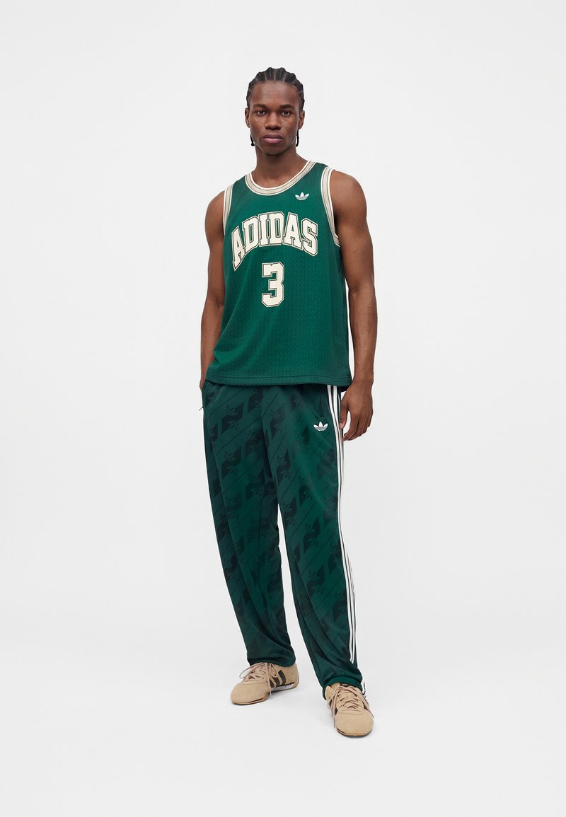 Mand stående iført grøn Adidas basketballtrøje med nummer 3 og matchende grønne træningsbukser med hvide striber og beige sneakers.
