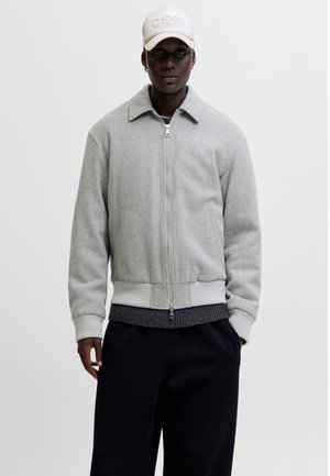 Homme portant une veste zippée gris clair, un pantalon noir, un pull foncé en dessous et une casquette blanche avec le texte « C1990 » sur le devant.