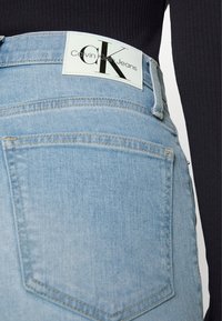 Ljusblå denimjeans med hög midja och bakfickor. Har en vit etikett med svart "CK"-logotyp. Bärs med en svart ribbad topp.