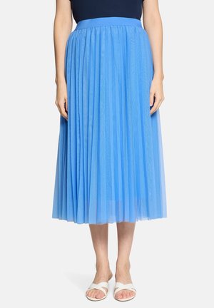 Frau trägt einen knielangen, hellblauen Faltenrock, dunkelblaues Oberteil und weiße Riemchensandalen, steht vor einem einfarbigen Hintergrund.