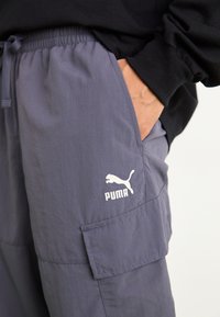 Graue Cargo-Hose aus Stoff mit elastischem Bund und Zugband. Enthält eine aufgesetzte Tasche und ein weißes Puma-Logo am Oberschenkel.