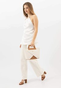Strukturalna torba z brązowym uchwytem, kremowym korpusem i kanciastym designem. Modelka nosi białą bluzkę i jasne lniane spodnie, stojąc w profilu.