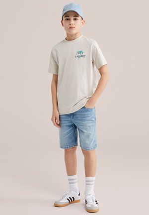 Niño con gorra azul claro, camiseta beige con la frase "Let's Explore", pantalones cortos de mezclilla, calcetas blancas a rayas y zapatillas blancas con rayas negras, de pie contra un fondo liso.