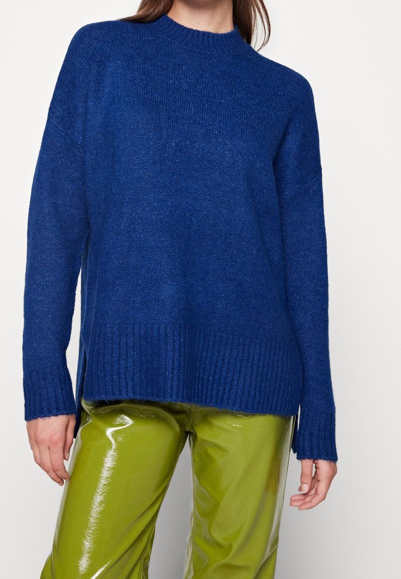 Femme portant un pull tricoté bleu avec des poignets et un ourlet côtelés, associé à un pantalon vert citron brillant sur un fond blanc uni.