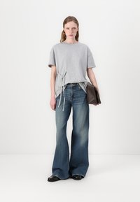 Šedé, přes oversized tričko s detailním uzlem po straně, spárované s rozšířenými modrými džínami. Modelka drží hnědou psaníčko. Jednoduchý design, volný střih.