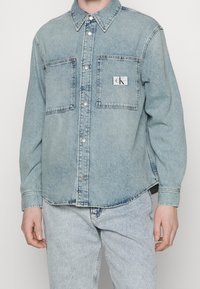 Camicia di jeans azzurro chiaro con colletto a punta, due tasche sul petto, bottoni a pressione in argento e patch con logo CK. Tessuto morbido e vestibilità comoda.