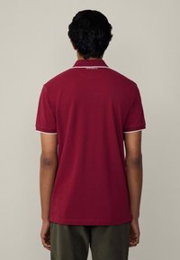 Bordová polokošeľa s klasickým golierom a bielym lemovaním pozdĺž rukávov. Na zadnej strane blízko krku sa nachádza malé logo.