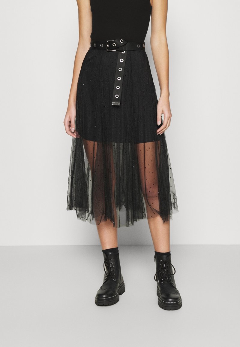 Jupe noire avec un voile en tulle transparent, texture à pois, et taille ceinturée avec des accents métalliques et un détail de boutons sur le devant. Bottes de combat noires.