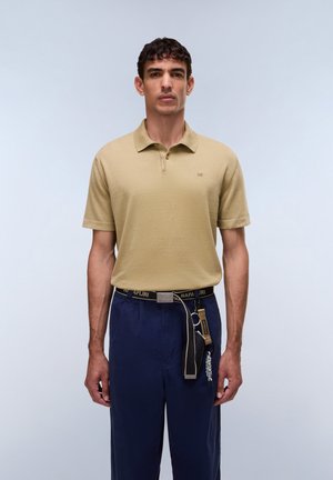 Homme portant un polo beige à manches courtes et un pantalon bleu marine avec ceinture noire de marque et accessoires porte-clés attachés, debout devant un fond uni.