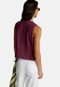 Kobieta z długimi brązowymi włosami ubrana w bezrękawnik w kolorze fioletowym, białe spodnie, okulary przeciwsłoneczne w postaci żółwi i trzyma zielono-biały szalik, stoi tyłem.