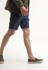 Farkkutrikoot tummansinisinä, käännetty reuna; yhdistetty oliivinvihreisiin liukuhyppykenkiin. Henkilö kesken askeleen, päällään tekstuurinen oliivinvihreä takki.