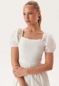 Witte jurk met korte pofmouwen, smock-borststuk en een vloeiende rok. De stof heeft subtiele texturen en patronen door het hele ontwerp.