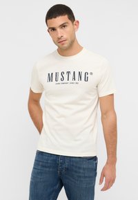 Kremowa bawełniana koszulka z krótkim rękawem, z wyrazistym granatowym logo "MUSTANG" i tekstem poniżej. Dżinsowe spodnie dopełniają stylizację.