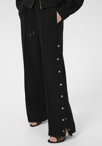 Schwarze weite Hosen mit seitlichen Taschen, Druckknopfdetails entlang der äußeren Nähte, gefertigt aus einem glatten Stoff und mit einem Kordelzugbund.
