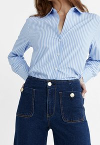 Mujer con camisa de botones a rayas azul claro y blanca, metida en unos pantalones vaqueros de tiro alto azul oscuro con grandes bolsillos frontales y botones dorados.