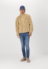 Chaqueta de fleece beige con cierre frontal, que presenta un pequeño parche con el logotipo. Combinada con jeans azules y zapatos beige, sobre un fondo sencillo.