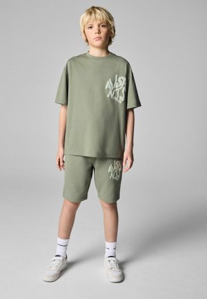 Šviesiai žalia medvilninė marškinėlių ir šortų komplektas su grafiniu atspaudų, laisvo silueto ir apvalia iškirpte. Dėvima su baltais sportbačiais ir kojinėmis.