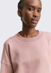 Sudadera de color rosa claro con cuello redondo, hombros caídos y una textura suave y lisa. Sin patrones ni acentos visibles.