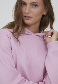 Helle rosa Kapuzenpullover aus weichem Material mit lockerer Passform. Verfügt über einen Kordelzug an der Kapuze und gerippte Bündchen. Das Modell hält den Stoff.