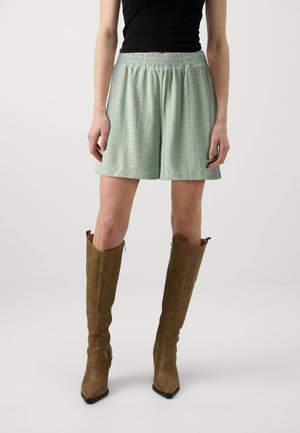 Shorts en lin vert clair avec un motif texturé, taille élastique et longueur jusqu'au milieu de la cuisse. Portés avec des bottes à talons haute jusqu'au genou marron.