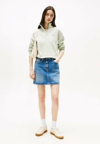 Sweatshirt verde chiaro cropped con mezza zip, abbinato a una gonna A-line in denim blu e sneakers bianche con suole beige. Borsa a tracolla semplice.