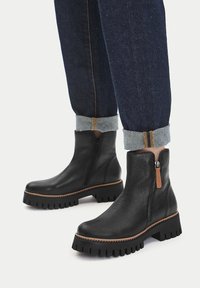 Bocanci negri din piele cu talpă groasă și fermoare laterale, purtați cu blugi denim întunecați, suflecați, pe un fond alb.