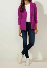 Fuchsiový blazer navrstvený přes bílý top, spárovaný s tmavě modrými džíny a bílými teniskami. Blazer má dlouhé rukávy a přiléhavý střih.