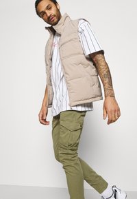 Beige Daunenweste mit hohem Kragen, zwei Vordertaschen; über einem gestreiften weißen Hemd getragen. Das Modell trägt olivfarbene Cargo-Hosen und Sneakers.