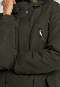 Olivgrön puffjacka med en foder av päls på huvan, dragkedjeförsedd bröstficka och elastiska ärmslut för värme och komfort.