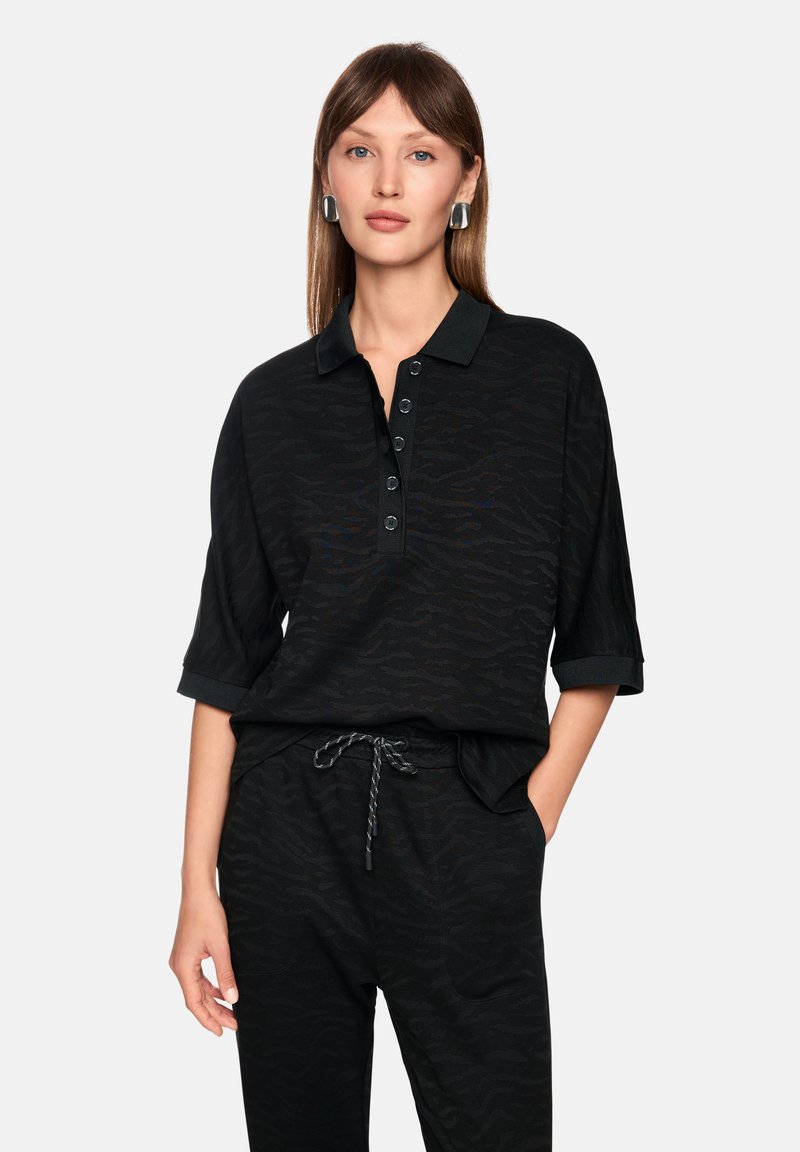 Schwarzes Poloshirt mit strukturiertem Zebramuster, halblangen Ärmeln, Knopfverschluss und kontrastierendem Kragen. Kombiniert mit passenden Hosen.