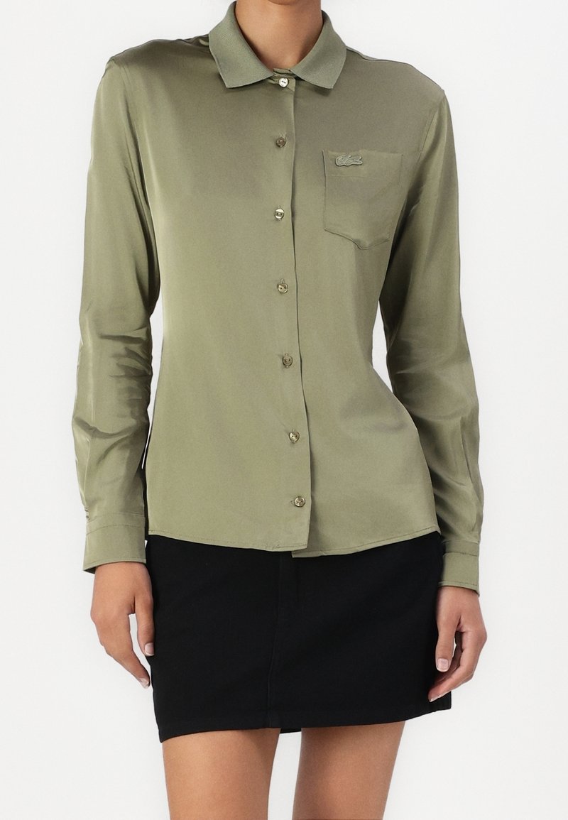 Femme portant une chemise à manches longues boutonnée vert olive avec une poche poitrine et une jupe noire sur un fond uni.