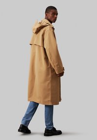 Beige vanntett trenchcoat med hette, med avslappet passform og sideslisser, kombinert med lyseblå jeans og svarte sko.
