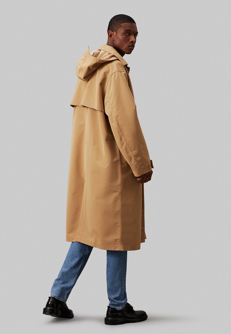 Beżowy płaszcz przeciwdeszczowy z kapturem, o luźnym kroju i bocznymi rozcięciami, zestawiony z jasnoniebieskimi dżinsami i czarnymi butami.