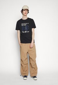 Zwarte katoenen T-shirt met witte en blauwe grafische tekst, gecombineerd met beige losvallende broek en marineblauwe sneakers met witte accenten, afgewerkt met een beige pet.