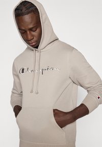 Μπεζ φούτερ με κουκούλα από βαμβάκι, με τσέπη μπροστά, κορδόνια στη κουκούλα και λαμπερό λογότυπο "Champion" στο στήθος.