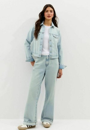 Vrouw draagt een lichtblauwe spijkerjas en wijde jeans over een wit T-shirt, staand tegen een effen witte achtergrond.