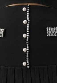 Vêtement en tissu noir orné d'une rangée d'accents perlés et d'un détail en perles le long du centre, avec une texture plissée en bas.