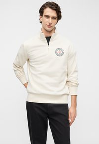 Κρεμ φούτερ με φερμουάρ και στρογγυλό λογότυπο στο στήθος, ριμπ μανσέτες και τελείωμα. Συνδυάζεται με σκούρο τζιν, με άνετη εφαρμογή και απαλό ύφασμα.