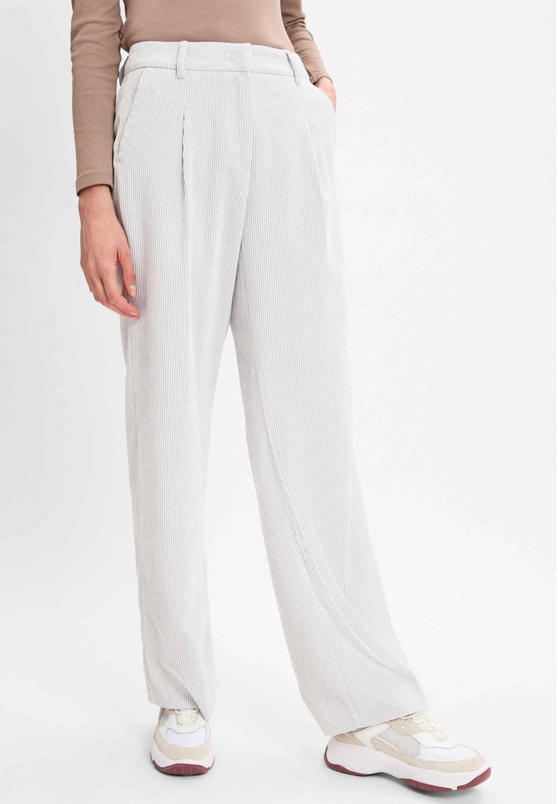 Personne portant un pantalon en velours côtelé gris clair taille haute à jambes larges avec des plis à l'avant et des baskets blanc beige, debout devant un fond blanc uni.
