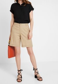 Black short-sleeve shirt, beige tailored shorts, and black strappy heeled sandals. Orange handbag adds a color accent.