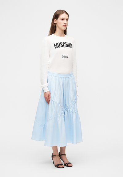 Γυναίκα με μακριά καστανά μαλλιά, που φοράει λευκό πουλόβερ Moschino Milano, φούστα μίντι ανοιχτού μπλε και μαύρες σανδάλες με ψηλό τακούνι, στέκεται στραμμένη προς τα δεξιά.