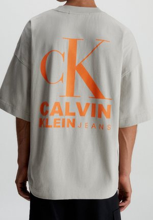 Light gray oversized t-shirt featuring bold orange "CK CALVIN KLEIN JEANS" text on the back. Made of soft cotton fabric.