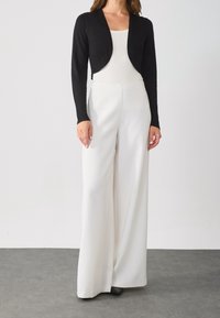 Black cropped cardigan over a white fitted top, paired with wide-leg white trousers. Smooth textures and minimal design with subtle seams.