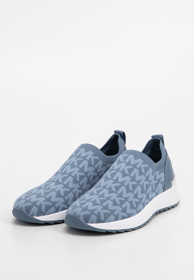 Des baskets slip-on bleues avec des semelles blanches et un tissu texturé présentant un motif géométrique répété, inclinées vers la gauche sur un fond blanc.