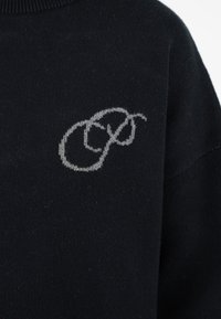 Schwarzer Strickpullover mit einem kleinen, hellgrauen gestickten Monogrammdesign im oberen Brustbereich.