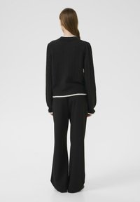 Pull en maille noire à manches longues, finition texturée et détail de ourlet blanc, associé à un pantalon noir ample. Vue arrière.